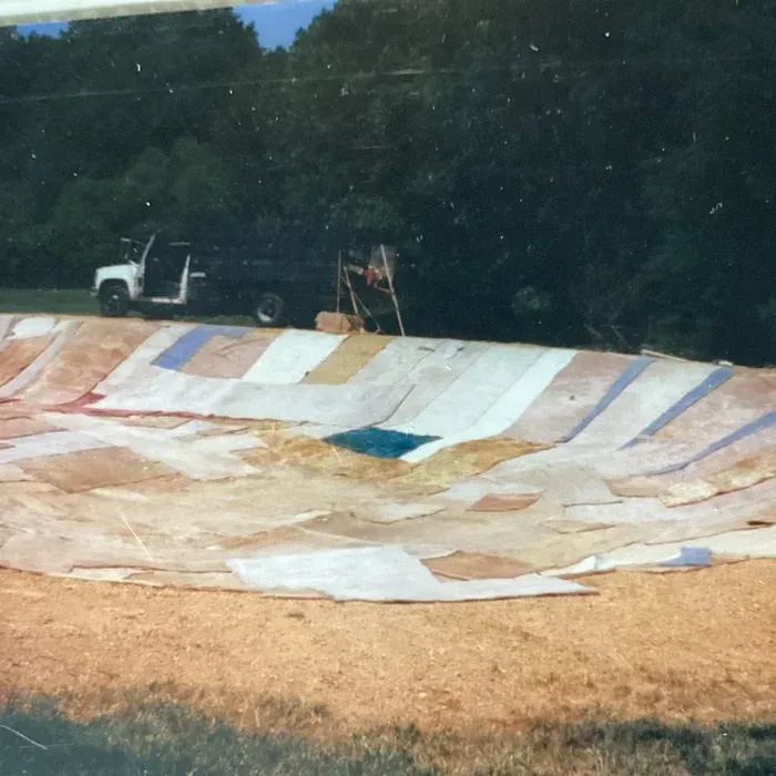 A large, shallow pit covered with a patchwork of white and tan fabric scraps sits before a truck near a wooded area.