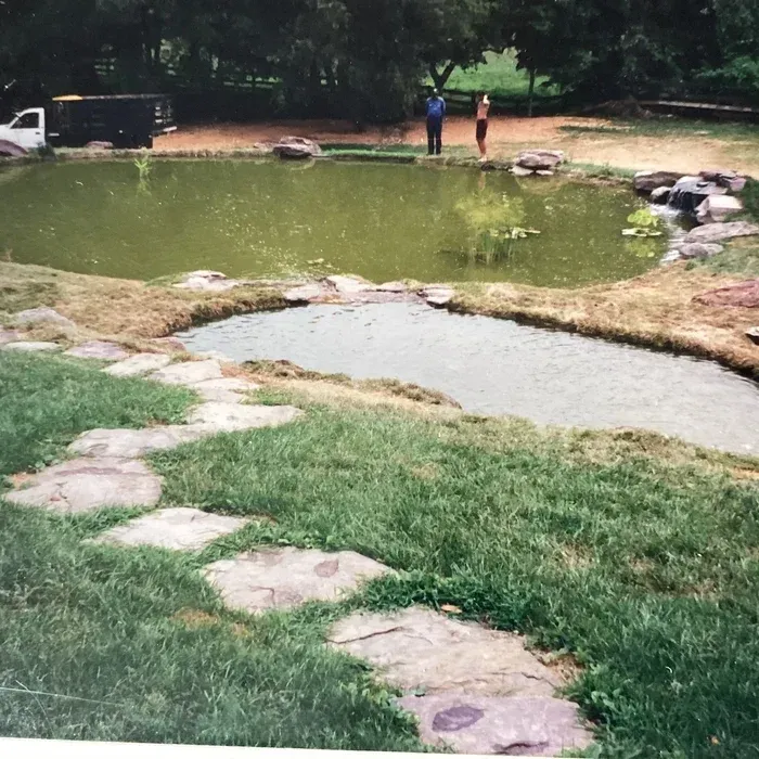 A stone path leads across a grassy lawn toward two connected ponds, with a vehicle and two people in the background.