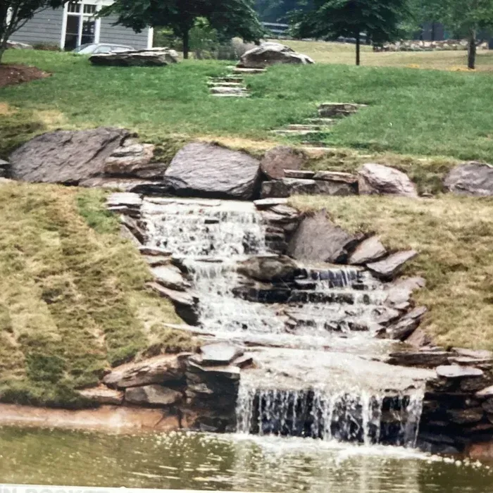 A tiered stone waterfall flows into a pond in a grassy yard, with stepping stones leading up the hill behind it.