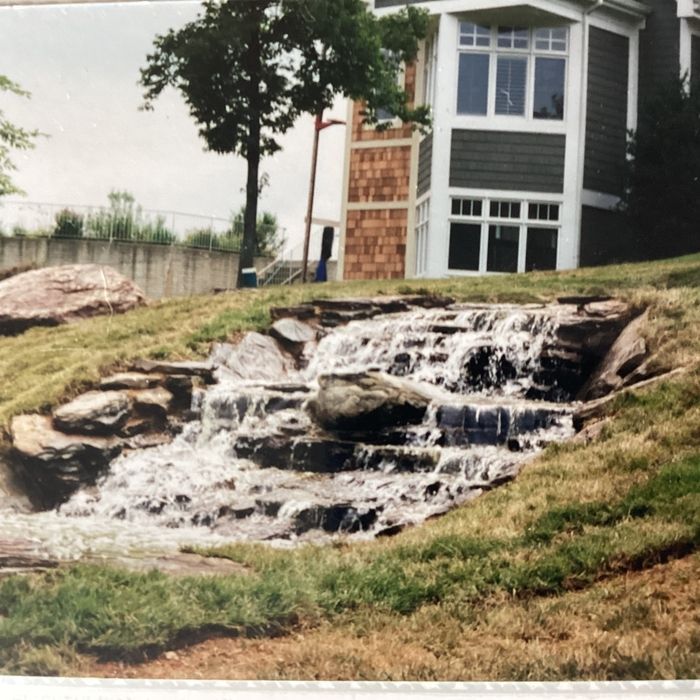 A man-made stone waterfall cascades down a grassy, sloped hill in front of a modern house with wood siding.