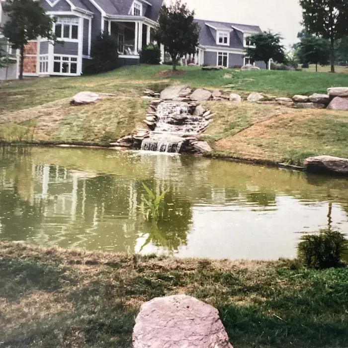 A backyard pond with a small rock waterfall, grassy lawn, and a multi-story house in the background.