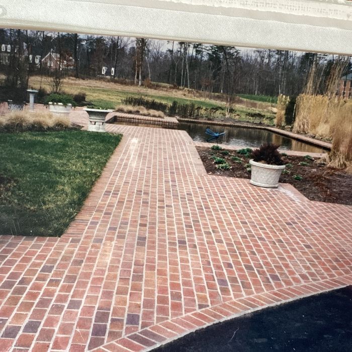 A red brick patio with a soldier course border extends toward a small pond in a landscaped garden.