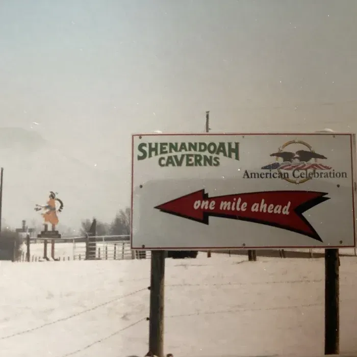 A road sign pointing left toward Shenandoah Caverns, set against a snow-covered field with a decorative figurine nearby.
