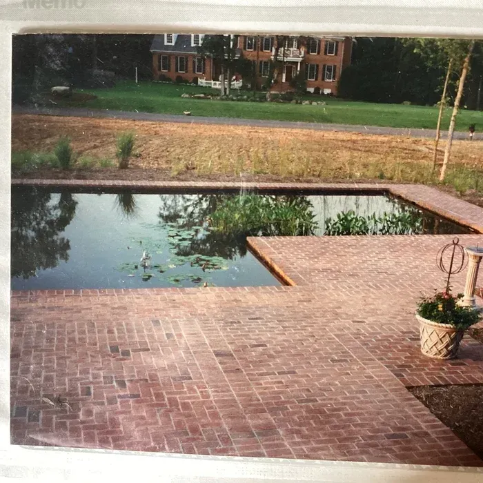 A rectangular pond surrounded by a red brick patio, with a brick house visible in the background.