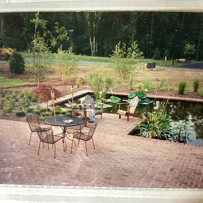 A patio with a round table, chairs, and a lounge chair sits beside a small pond in a grassy yard backed by trees.