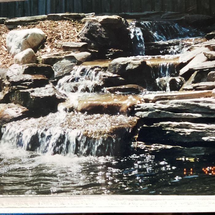A multi-tiered stone waterfall cascades into a dark pond, with layers of rock and scattered mulch in the background.