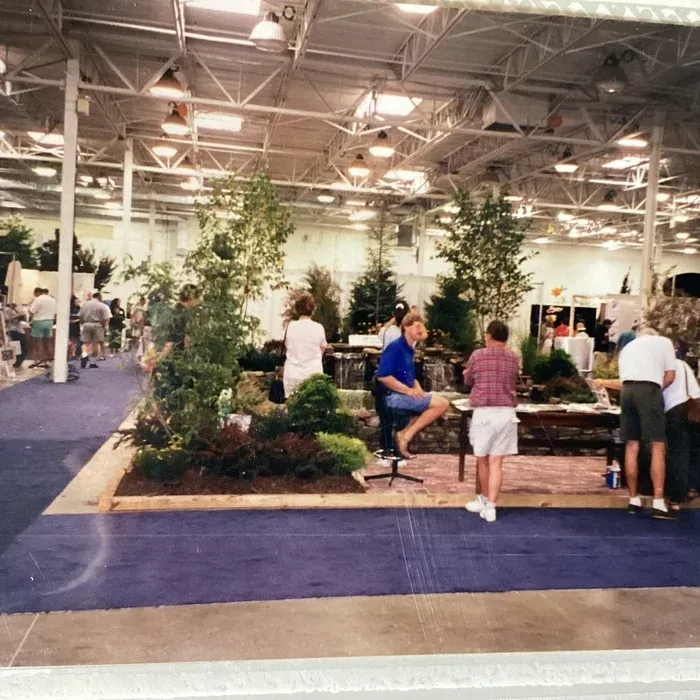 People explore a landscaping and water feature display at an indoor exhibition hall with blue carpet and metal ceilings.