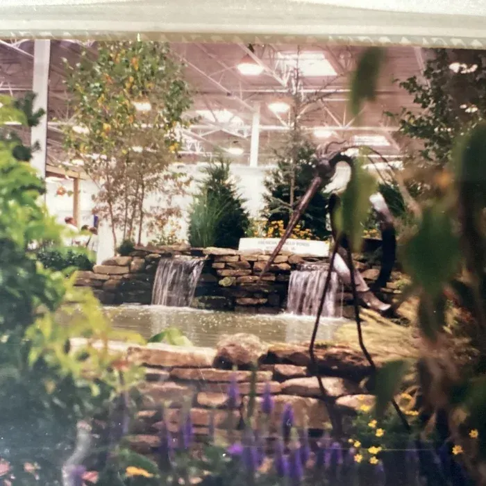 A tiered stone waterfall flows into a pond in an indoor garden display, with a metal bird sculpture standing nearby.