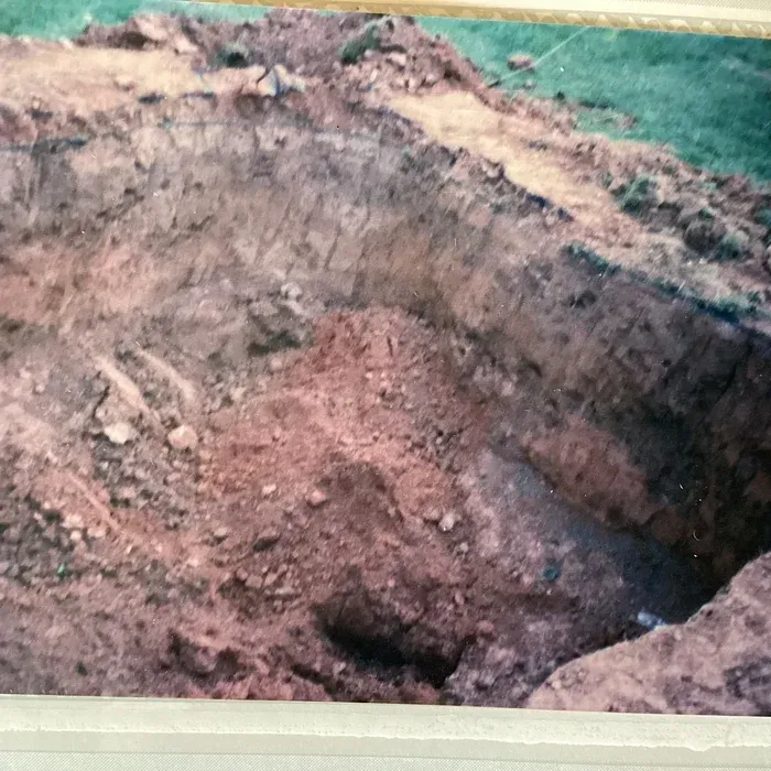A deep, rectangular earthen pit dug into reddish-brown soil, surrounded by patches of grass.