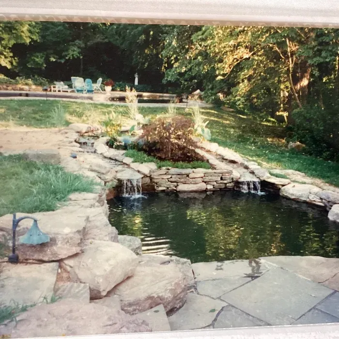 A stone-lined koi pond with two small waterfalls and surrounding landscaping in a grassy yard.