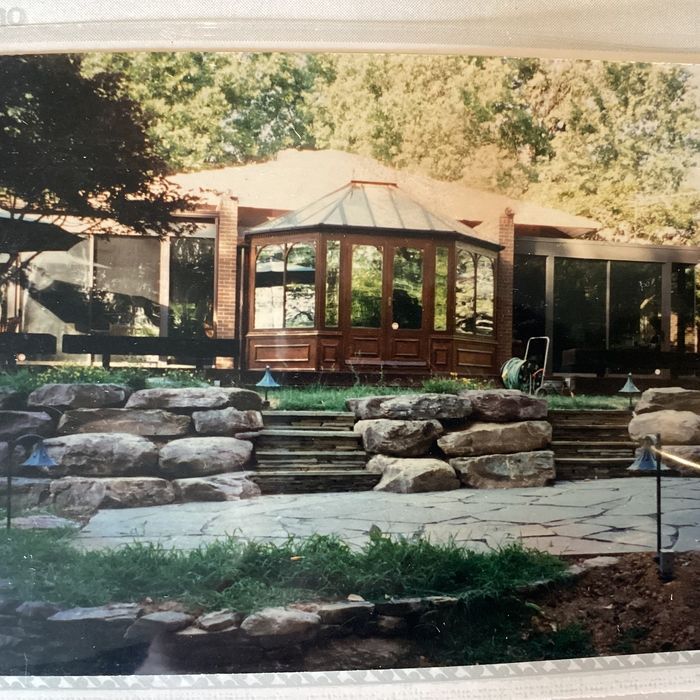 A backyard scene featuring a home with a central wooden sunroom, a flagstone patio, and a stone retaining wall with steps.