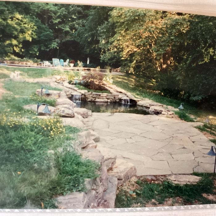 A stone patio overlooks a small pond with a waterfall, surrounded by lush green trees and landscaping.