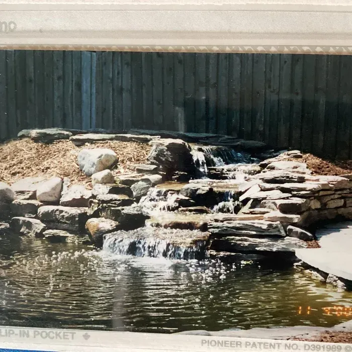 A cascading stone waterfall flows into a pond in a backyard setting in front of a wooden fence.