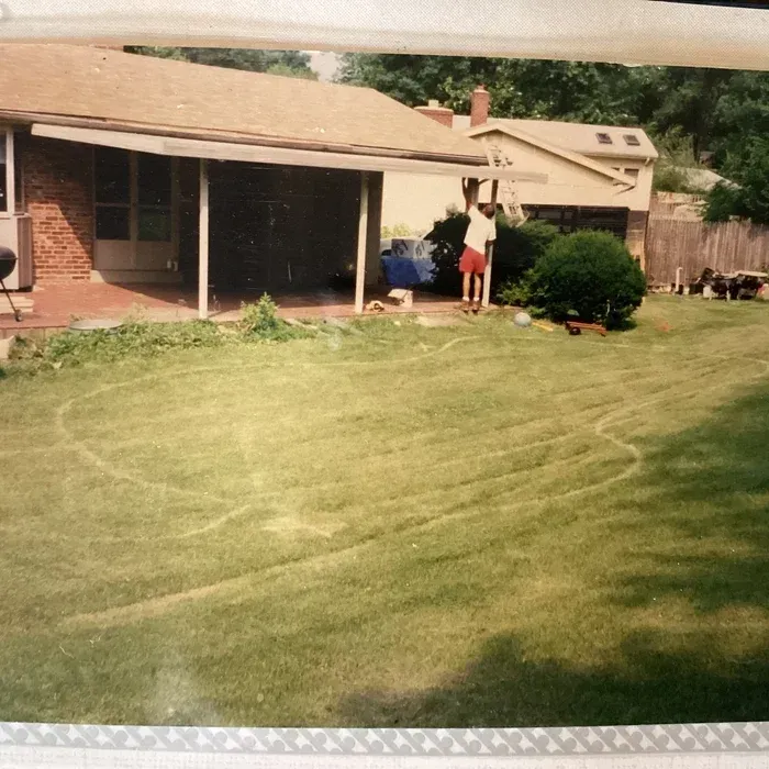 A person stands under a patio roof in a backyard with a large, circular chalk outline drawn on the green lawn.