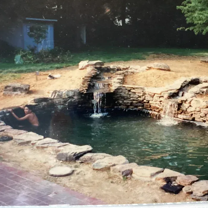 A person in a backyard pond with two small waterfalls cascading over stone rocks into the water.