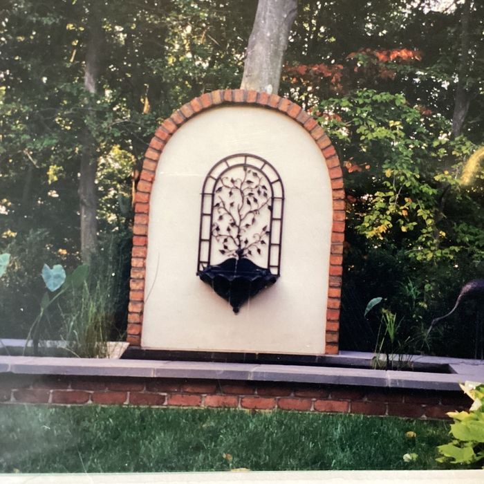 A brick-arched fountain wall featuring a metal lattice tree sculpture, set in a garden with greenery.