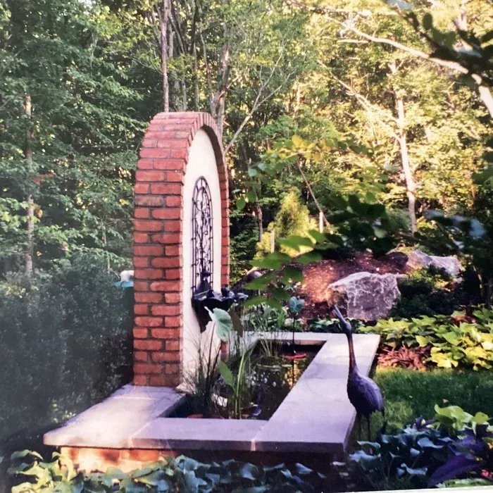 A brick arch fountain and rectangular stone pond sit in a wooded garden, with a decorative metal heron sculpture nearby.