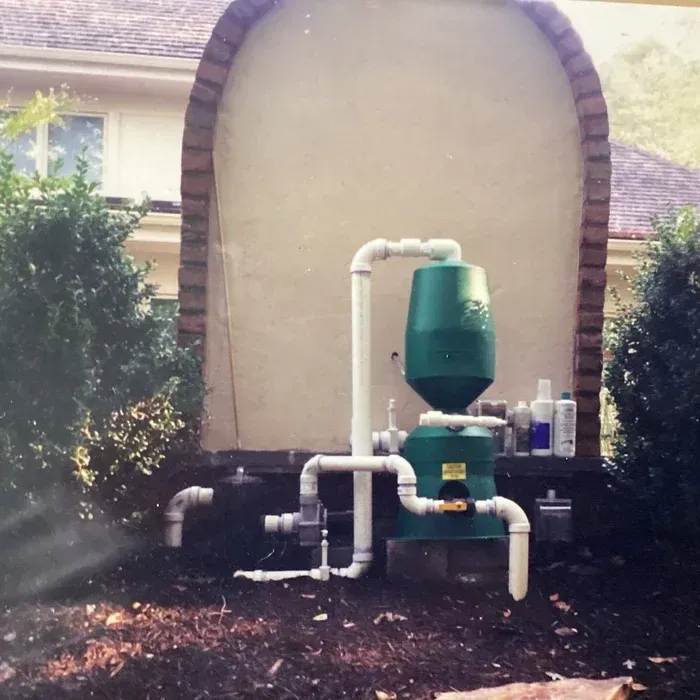 A green pool filtration system with white PVC pipes is installed outdoors against a tan wall between two bushes.