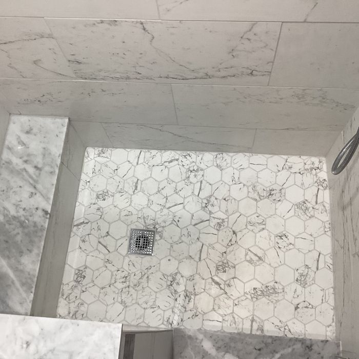 A high-angle view of a tiled shower floor with white, marble-patterned hexagonal tiles and a center metal drain.