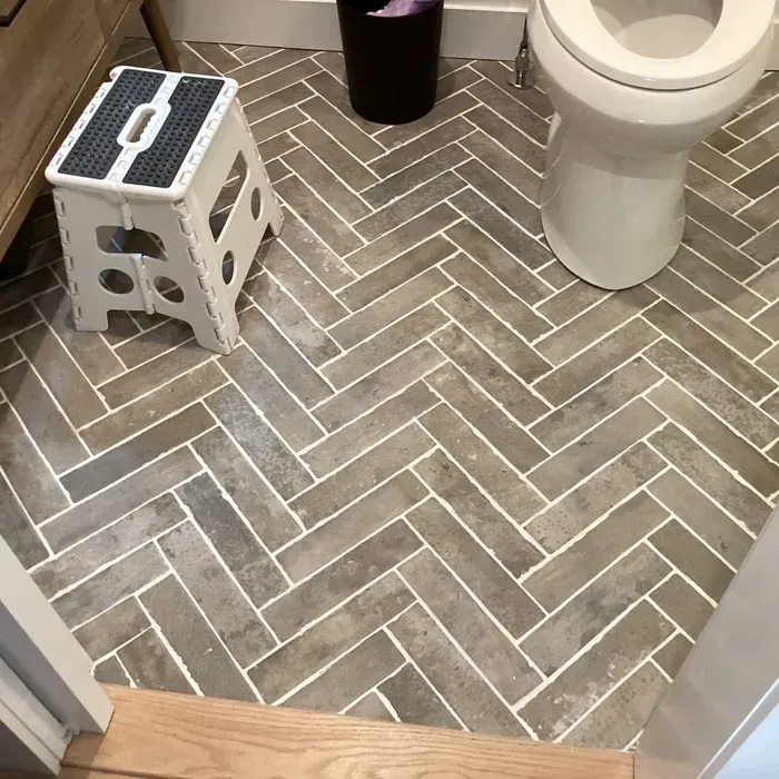 A bathroom floor with light-grey brick-style tiles in a herringbone pattern, featuring a white step stool and a toilet.