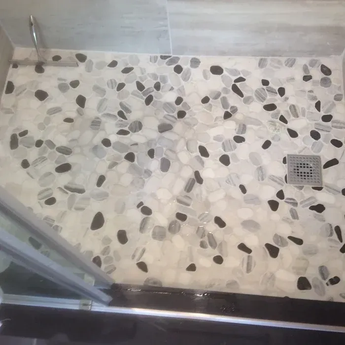 A modern shower floor featuring a pebble mosaic tile pattern in white, gray, and black, with a square metal drain.