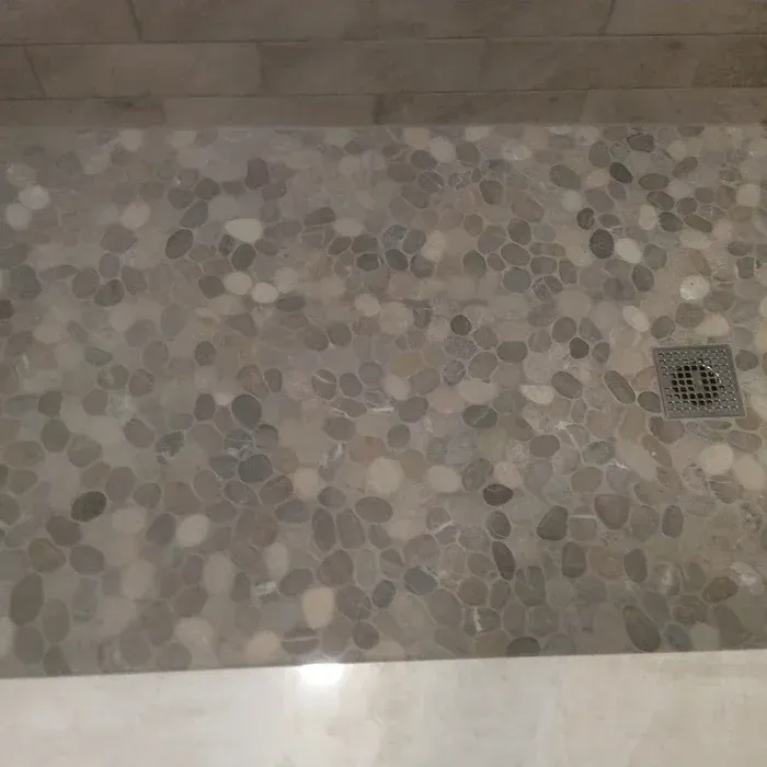 A shower floor made of smooth, grey and tan pebble tiles, featuring a square metal drain on the right.