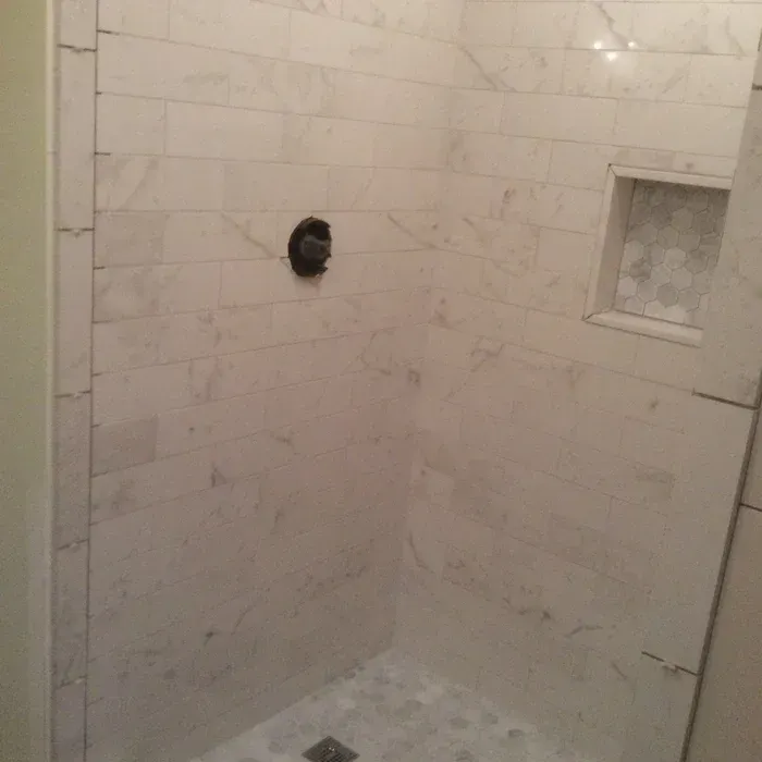 A shower interior with white marble wall tiles, a black circular faucet handle, a square recessed niche, and a tiled floor.
