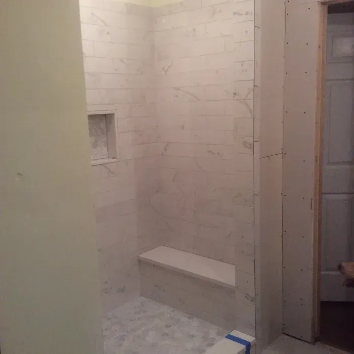 A partially tiled shower featuring light marble wall tiles, a built-in bench, a small niche, and a stone pebble floor.