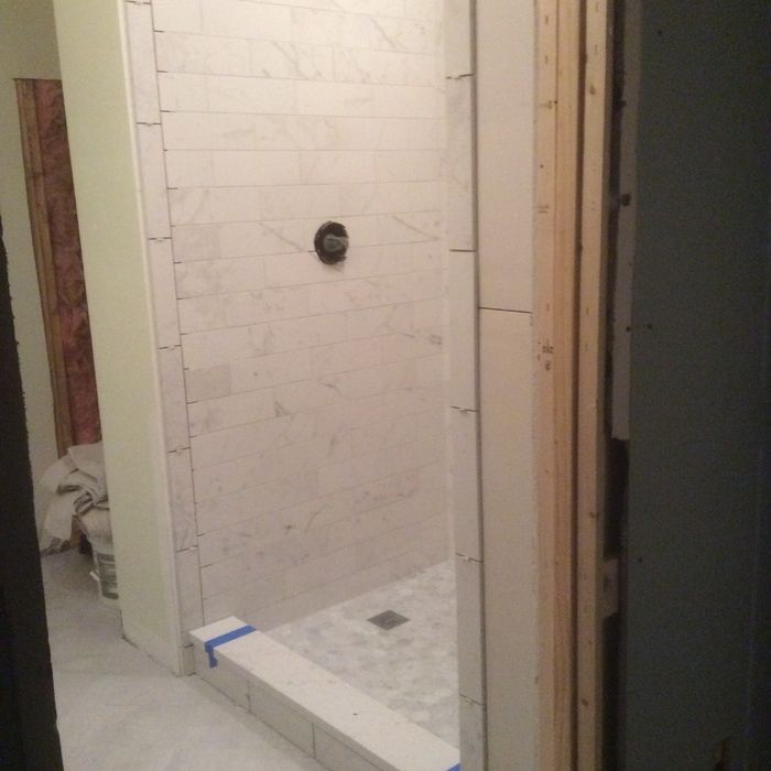 A partially completed walk-in shower with light-colored tile walls, a dark shower handle, and a tiled curb.