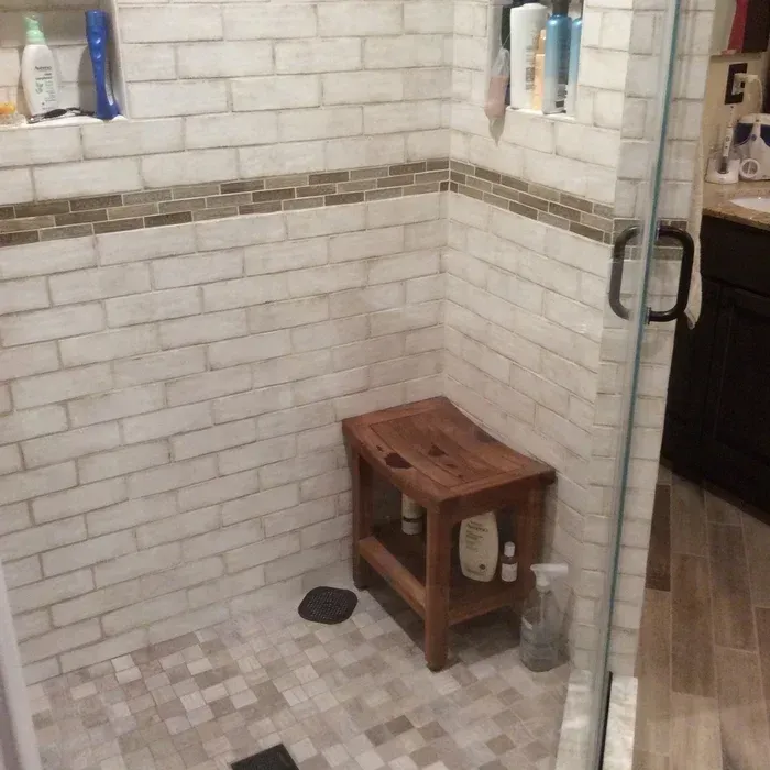 A walk-in shower with white brick-style walls, a brown mosaic tile border, and a wooden stool placed in the corner.