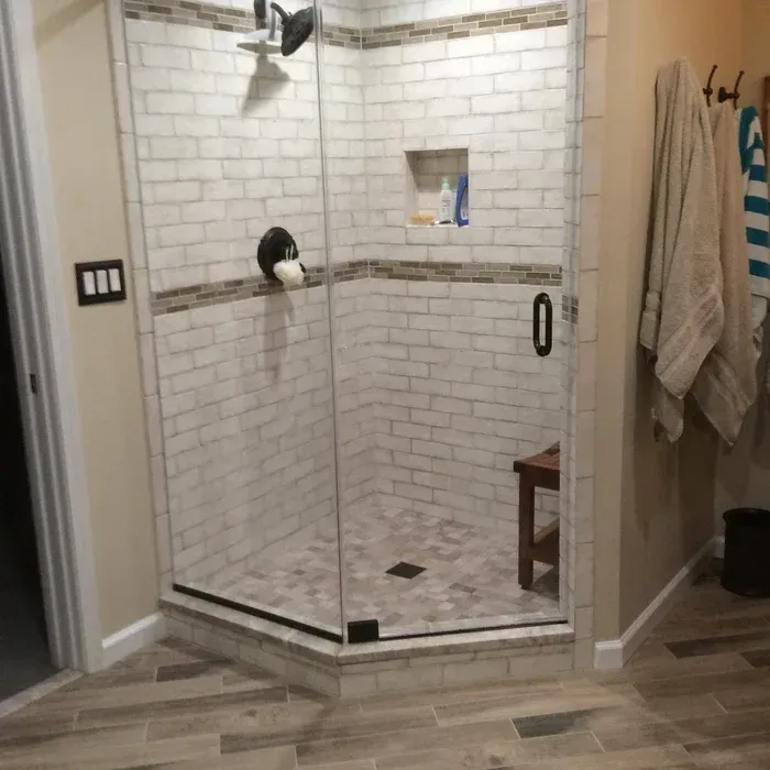 A corner glass shower stall with white brick-patterned tiles, a decorative accent band, and a small wooden bench inside.