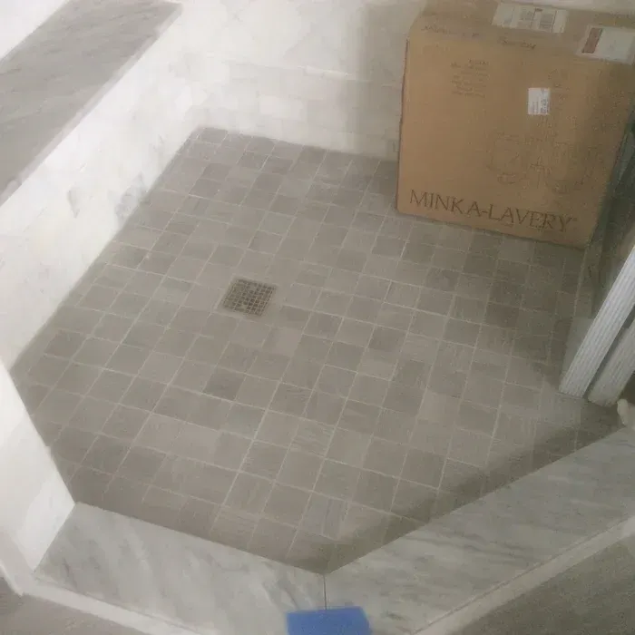A tiled shower floor with a central drain, next to a cardboard box and a marble-tiled curb in a room under construction.