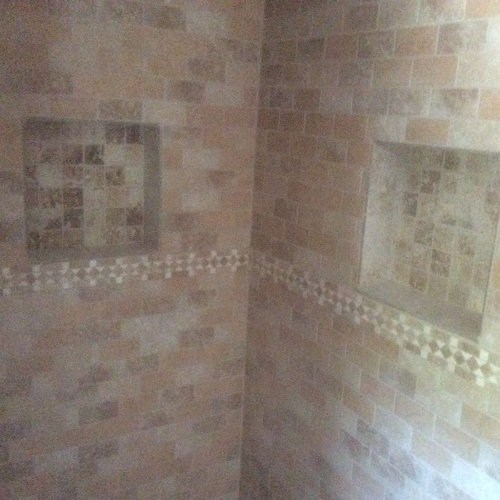 Shower wall with beige rectangular tiles, a decorative mosaic border, and two inset wall niches with square stone tiles.