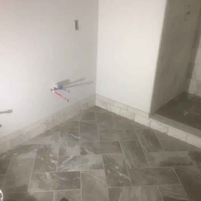 A corner in a room featuring gray herringbone-patterned tile flooring, a stone baseboard, and exposed plumbing pipes.