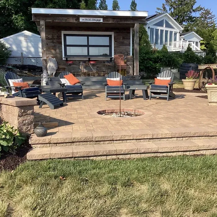 A stone patio features a fire pit, several outdoor chairs with orange cushions, and a rustic wooden covered bar area.