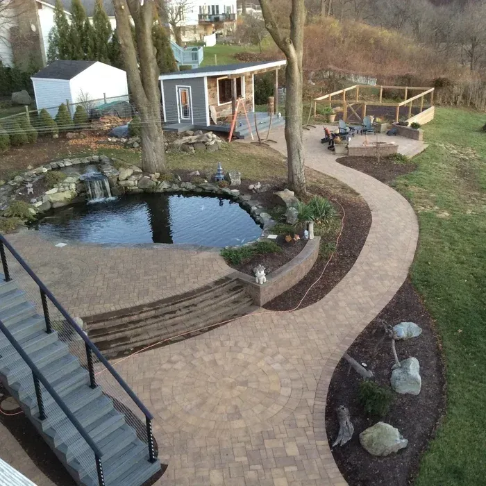 A landscaped backyard features a pond with a waterfall, a stone patio, a curved walkway, and a small wooden structure.