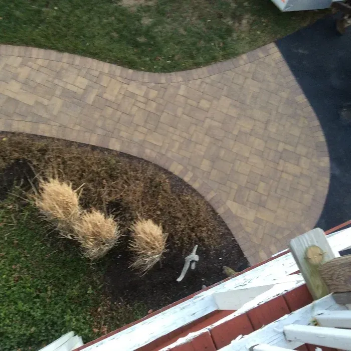 A curved stone paver walkway meets a black driveway, viewed from above near a wooden deck with railings.