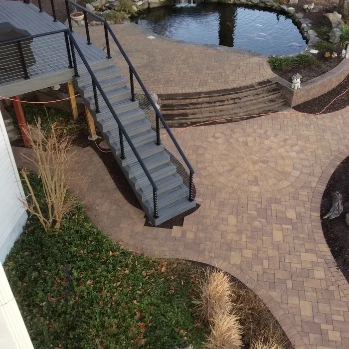 A gray deck staircase leads down to a patterned stone patio and a curved backyard pond in a landscape setting.