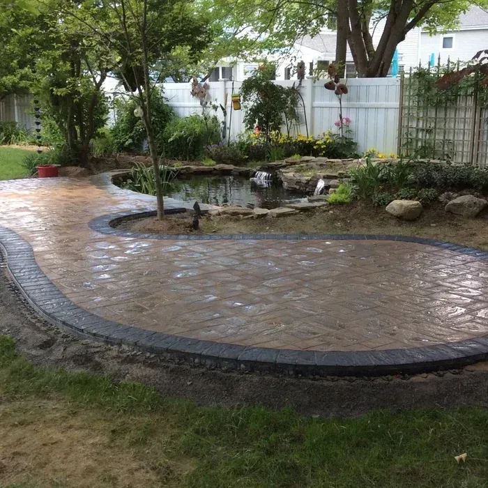 A curved stone patio with a dark paver border sits next to a small garden pond in a grassy backyard.