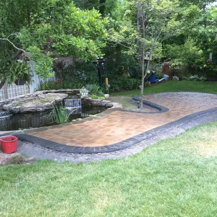 A landscaped backyard with a stone patio, a small rock waterfall pond, and lush green trees in a sunny garden setting.