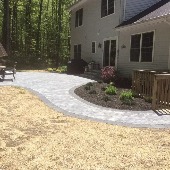A light-colored stone patio curves past a mulched garden bed next to the gray siding of a two-story house.