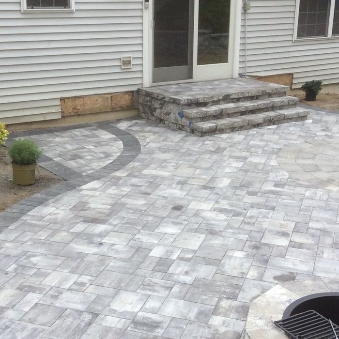 A light grey stone patio with a curved dark border, leading up to stone steps against a light siding house exterior.