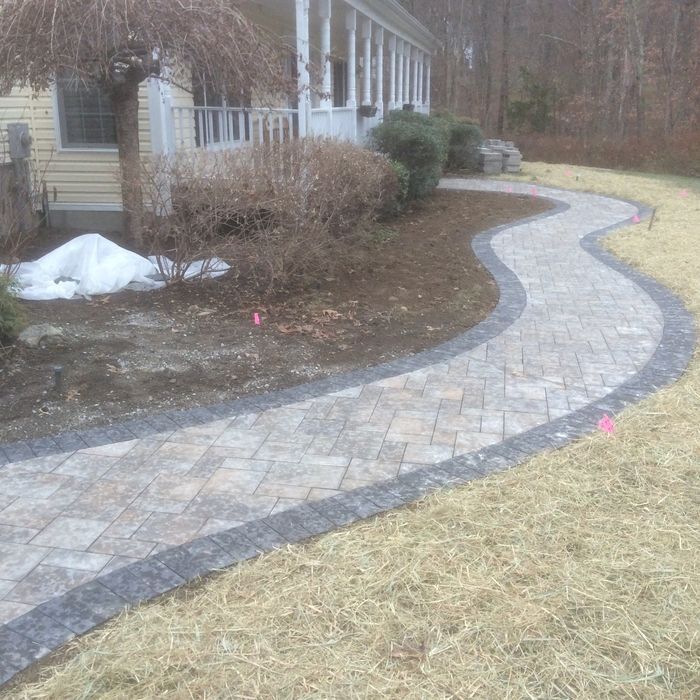 A curved paver walkway with a dark border leads toward a house with a porch and landscaping in a residential yard.