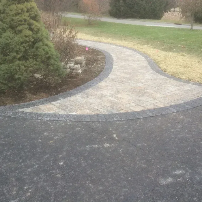 A paved walkway with a tan stone center and dark stone border transitions from a black asphalt driveway to a lawn area.