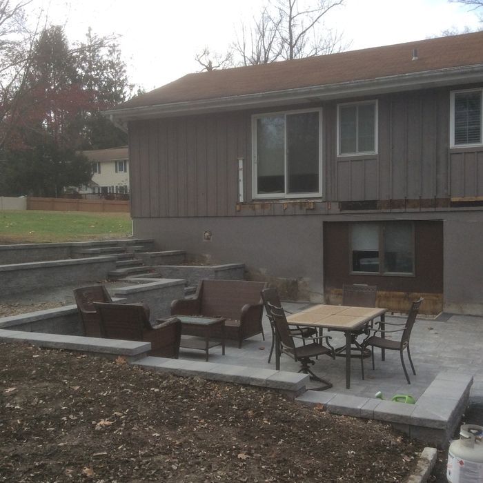A backyard patio with a dining table, chairs, and bench, set against a two-story house with tiered retaining walls.
