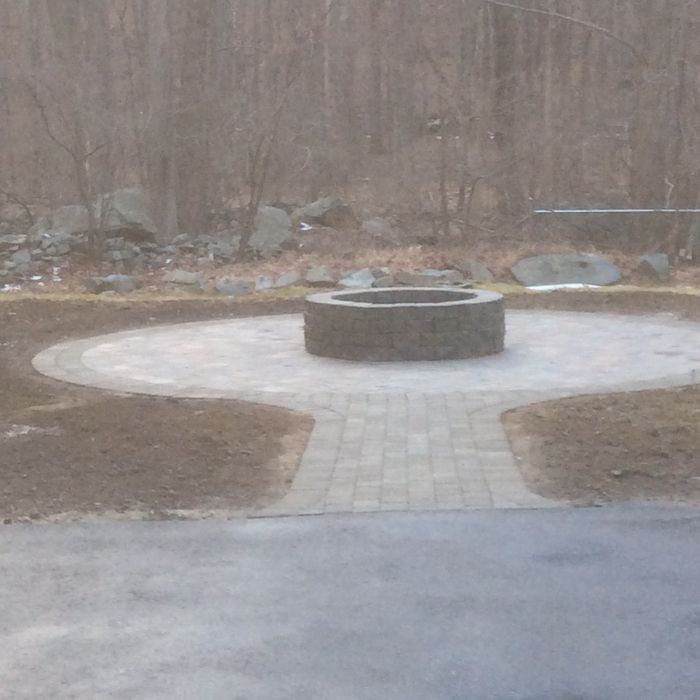 A circular stone patio with a central fire pit, connected to a paved driveway by a straight stone walkway in a yard.