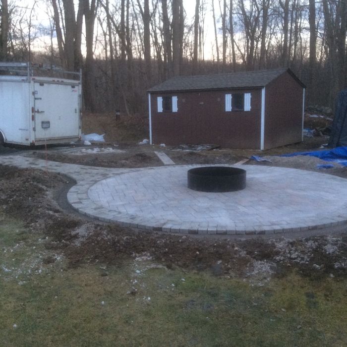 A circular stone patio with a central fire pit sits in a yard near a small shed and a white trailer.