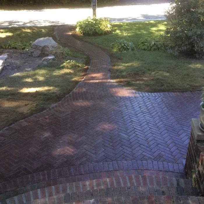 A herringbone brick path winds through a lawn, leading from a brick porch toward a driveway with a large boulder nearby.