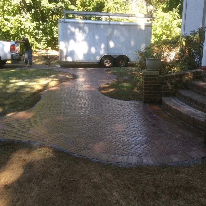 A person stands near a white utility trailer parked on a lawn next to a newly paved herringbone brick walkway and steps.