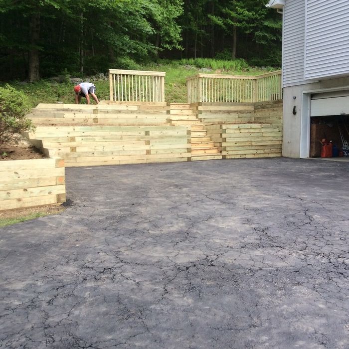 A person building tiered landscape retaining walls made of wooden timbers next to a paved driveway and garage.
